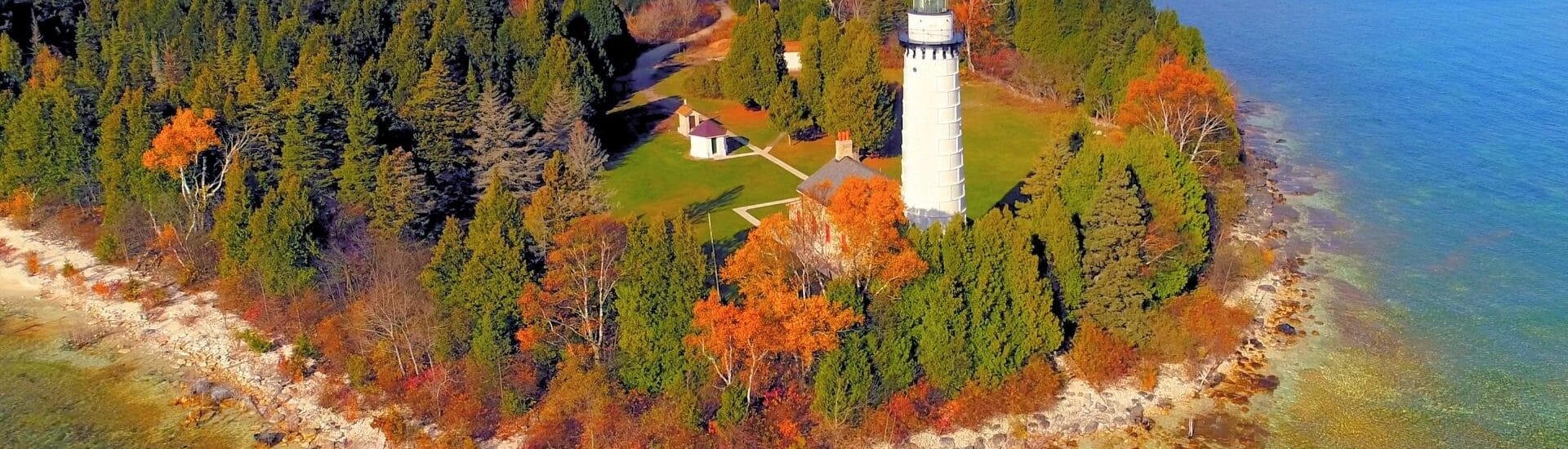 aerial view of a lighthouse in door county surrounded by fall trees on a rocky shoreline with blue lake water