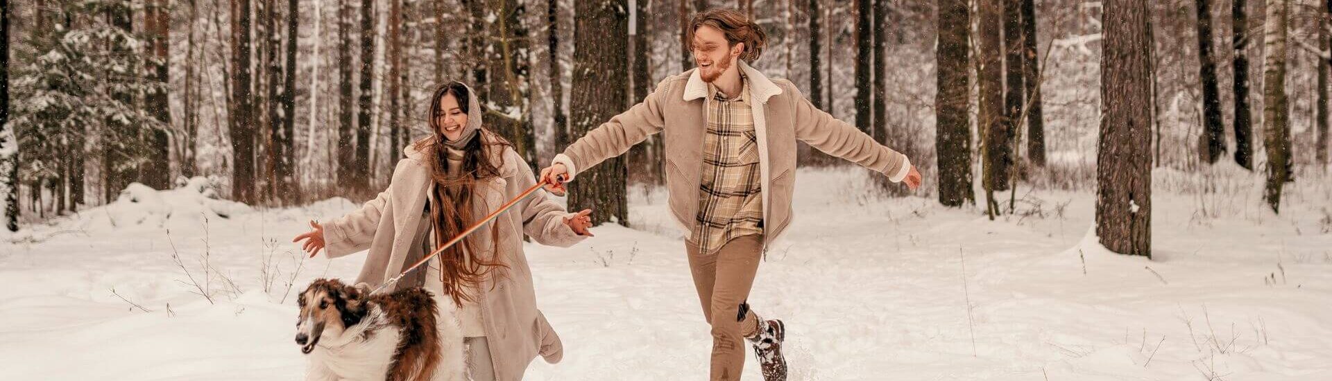 smiling couple in beige coats runs through the snow with their dog surrounded by tall trees