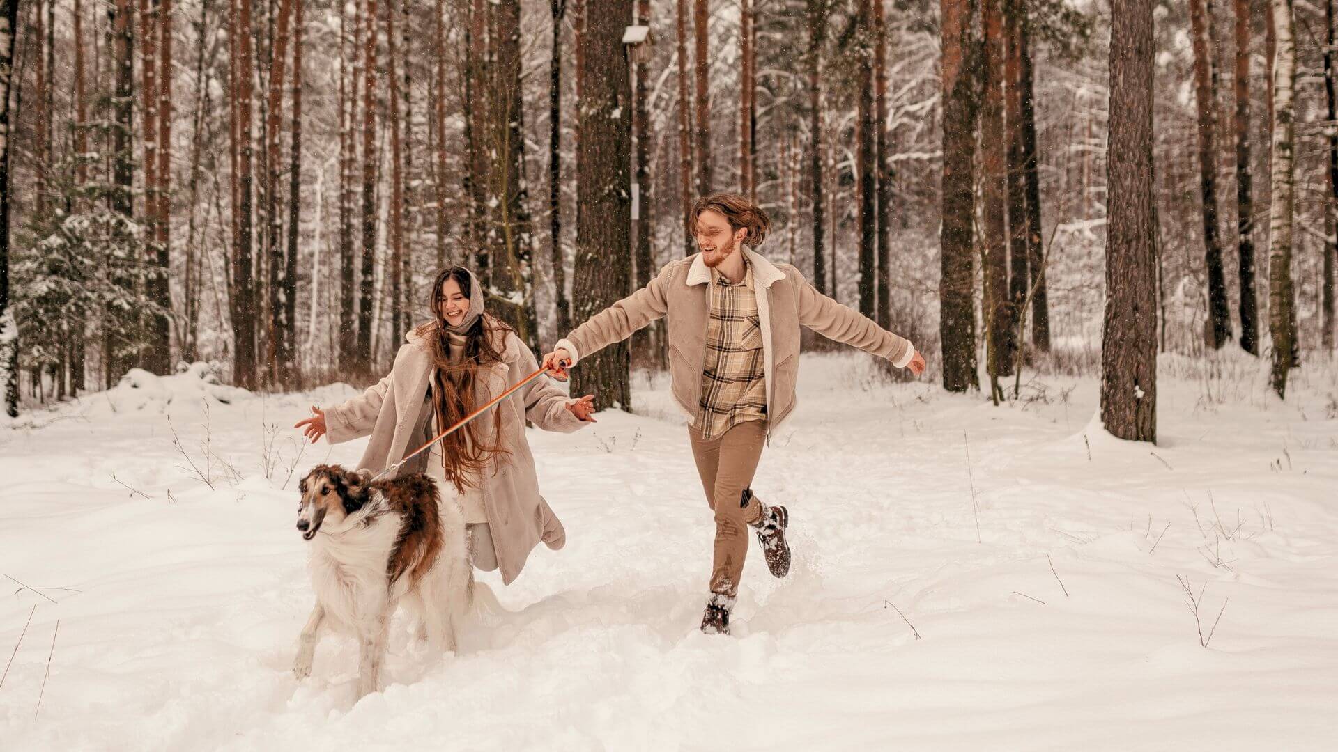smiling couple in beige coats runs through the snow with their dog surrounded by tall trees