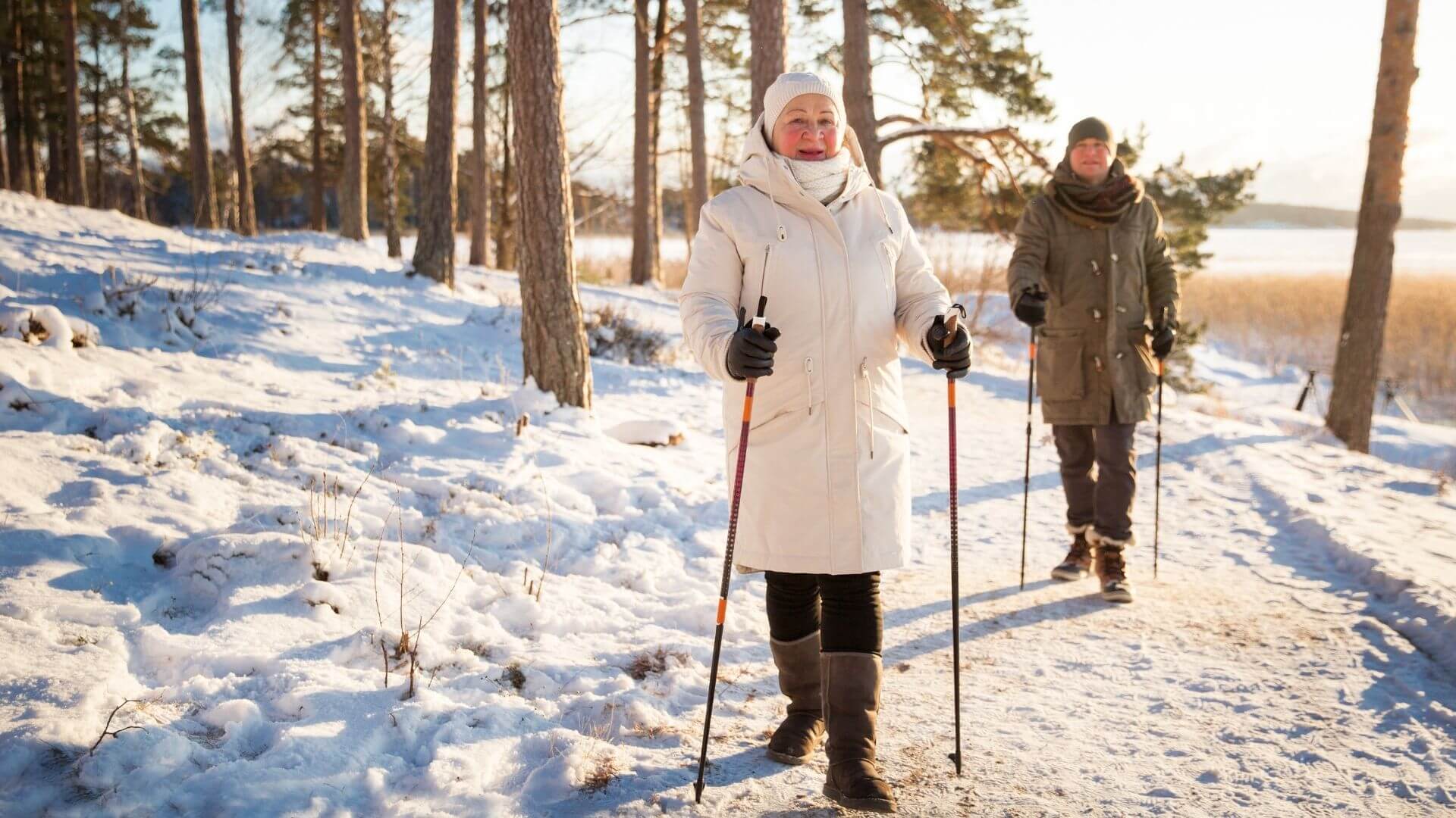 two people dressed in warm coats and hats use walking poles along a snowy forest trail beside a frozen lake