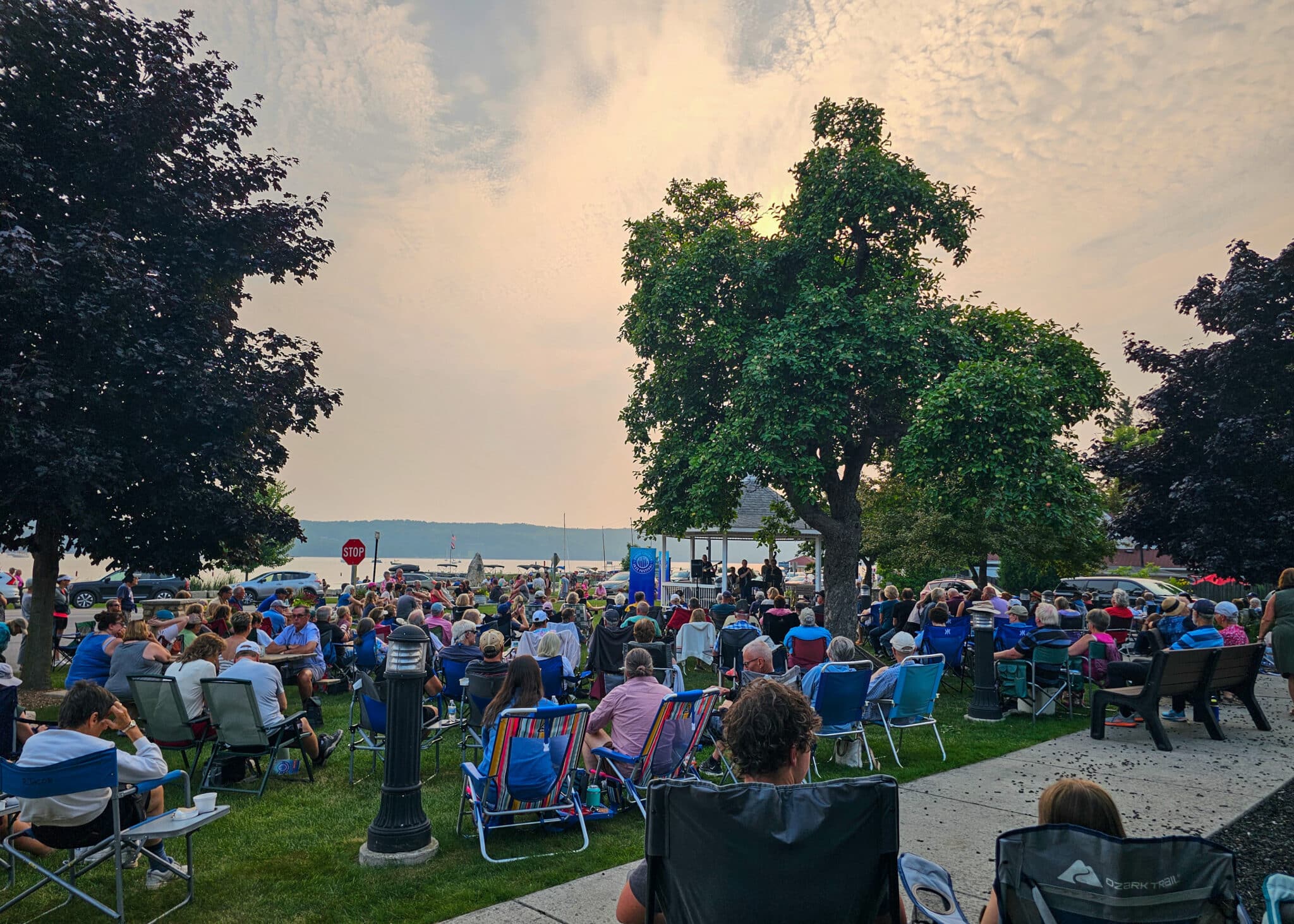 Large crowd seated on folding chairs in a lakeside park, watching a distant stage at sunset/late afternoon light.