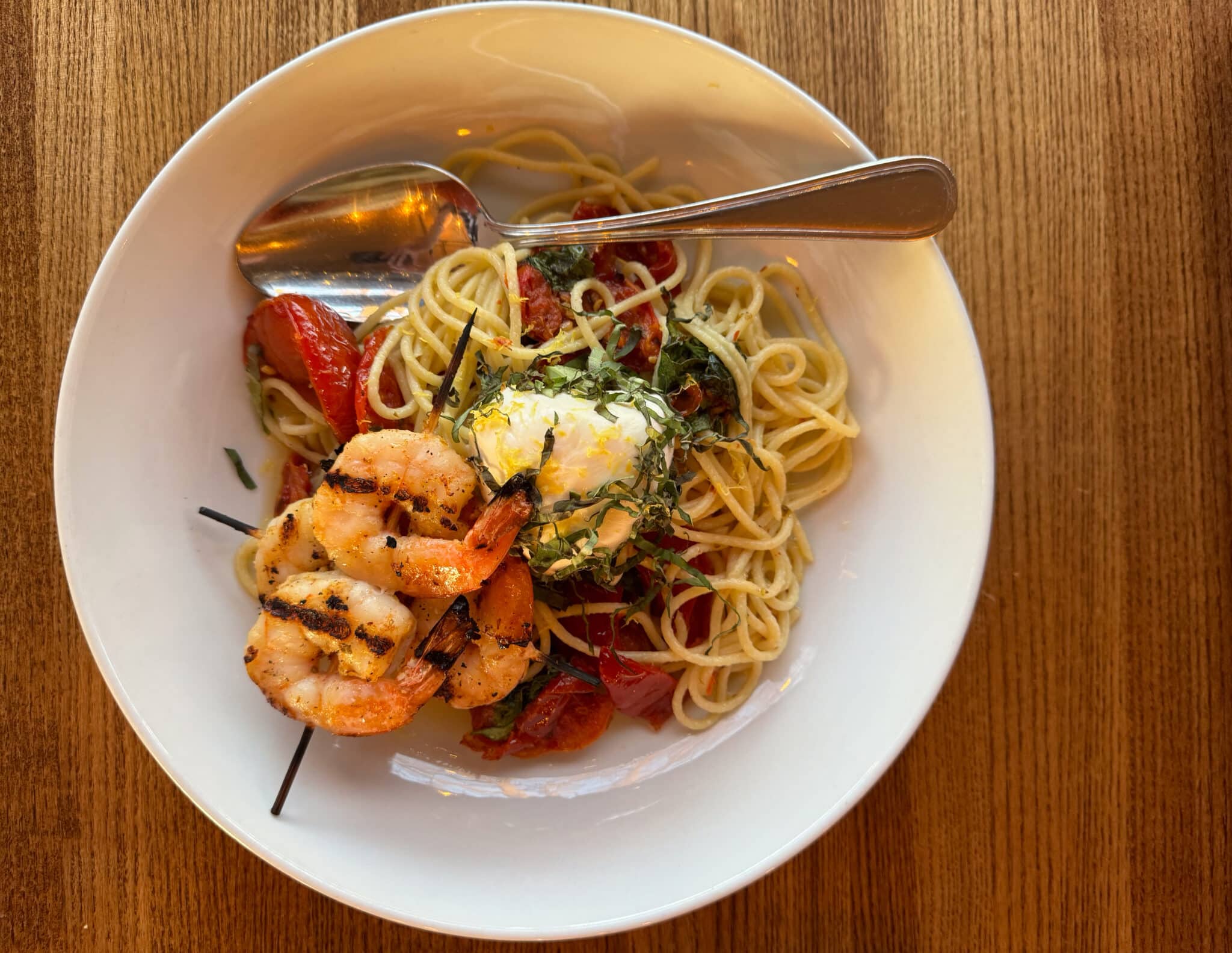 Spaghetti with grilled shrimp, cherry tomatoes, herbs, and lemon zest on a white plate set on a wooden table, with a spoon across the pasta