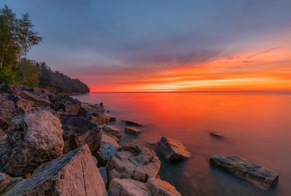 Rocky shoreline with trees on the bluff and a gorgeous sunset sky with bright orange and yellow colors