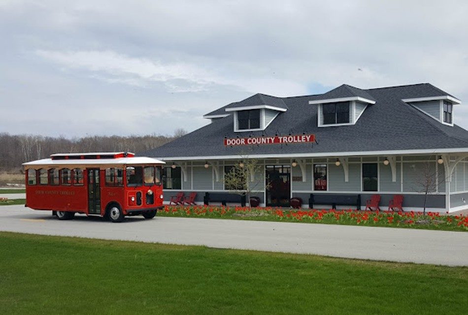 A red trolley sitting on a paved road next to a large grey building