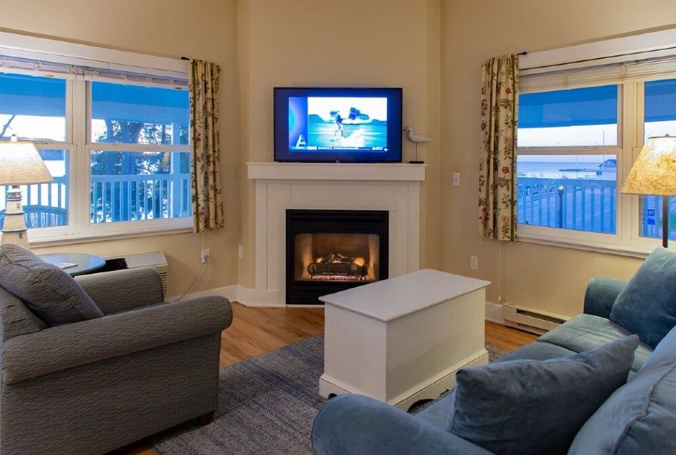 Cozy living room with plush couch and chair, table, gas fireplace with TV and two large windows with floral curtains