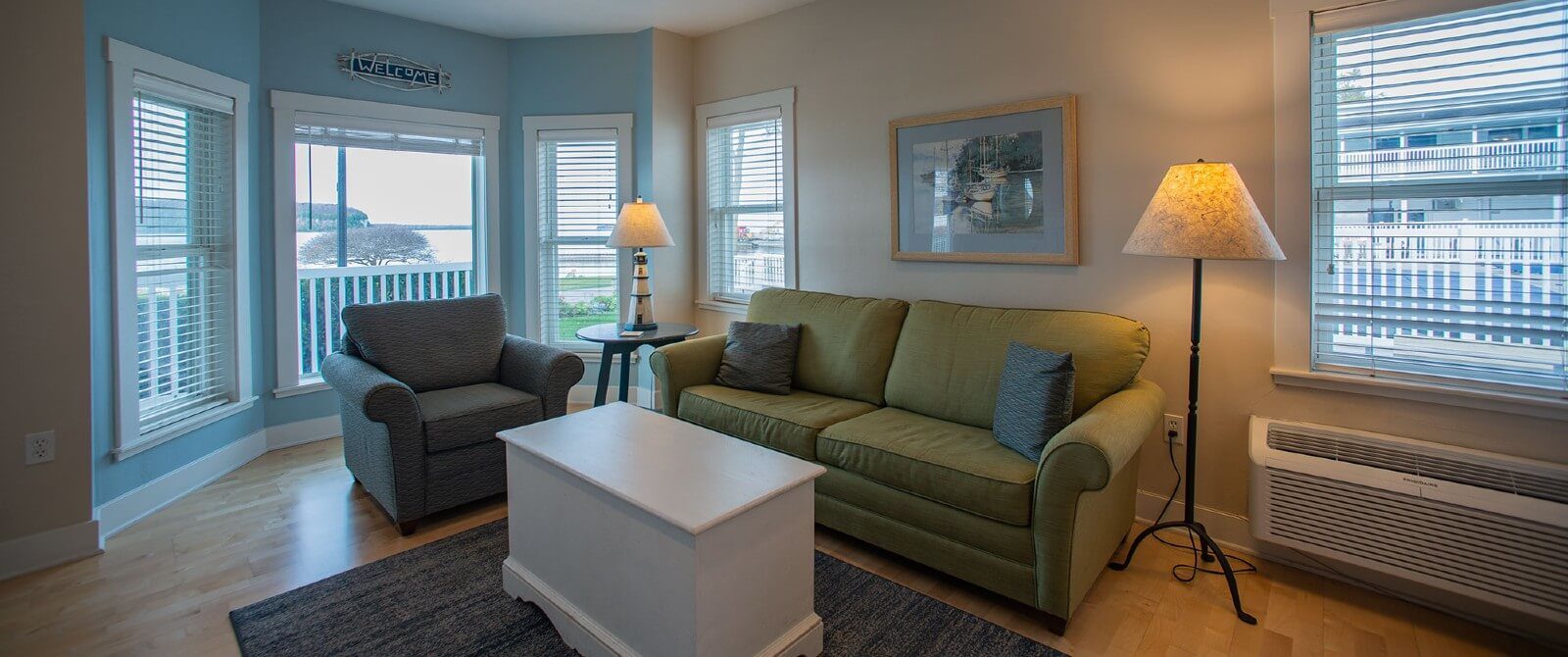 Living room area of a suite with two lamps, a green couch, chair, table and bay windows overlooking water