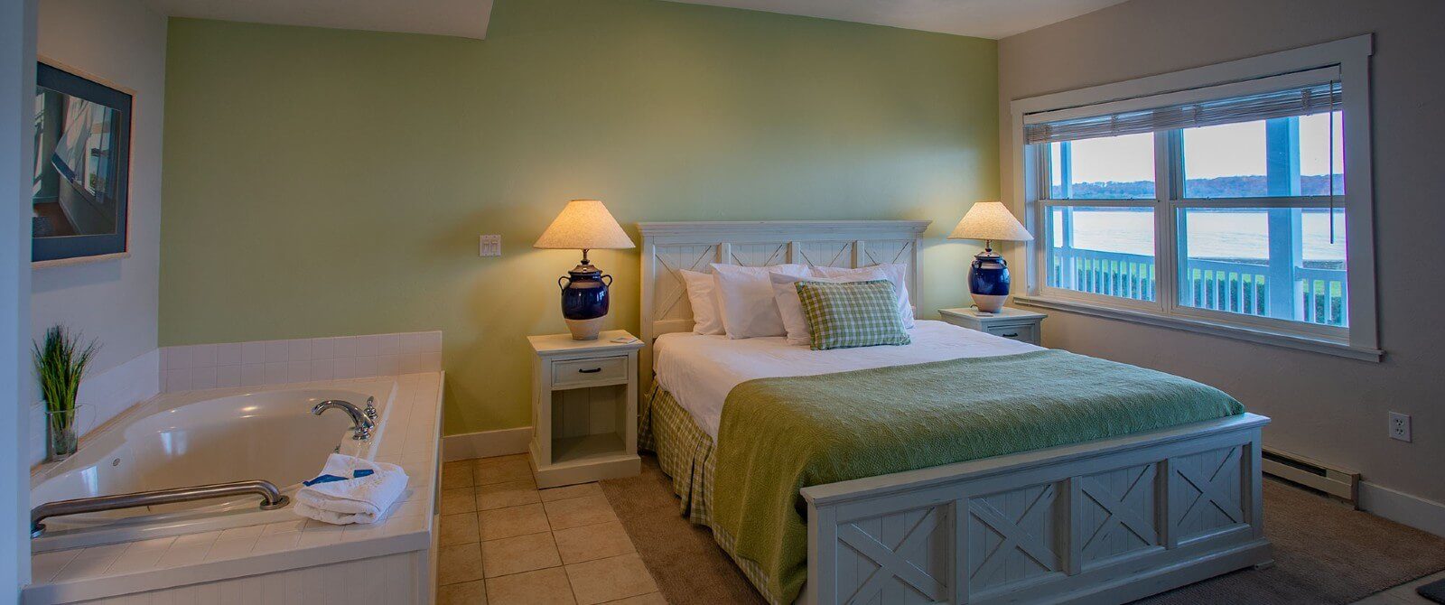 Bedroom with queen bed by a large window, tables with lamps and jacuzzi tub