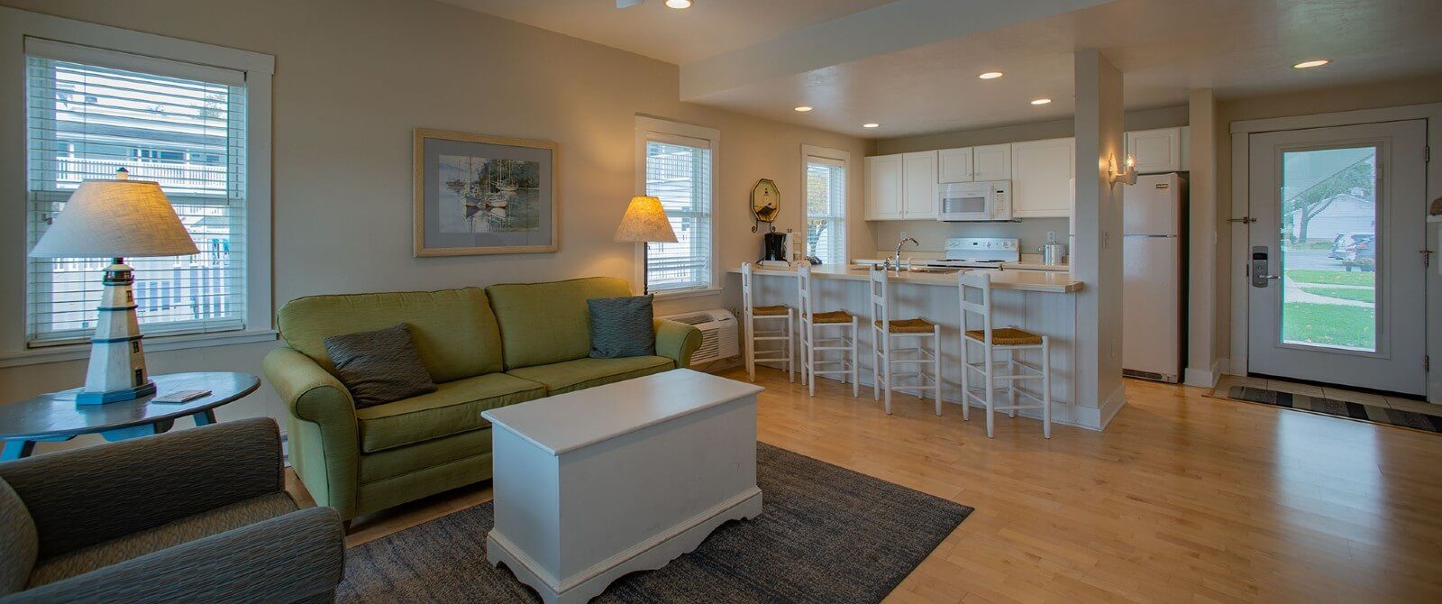 Large hotel suite with hardwood floors, full white kitchen, bar stools, living room with couch, chair and white table on an area rug.