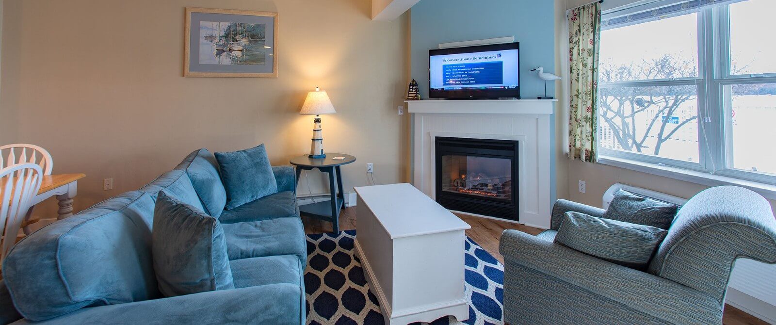 Living area in a suite with large window, blue couch and chair, table and TV over a gas fireplace