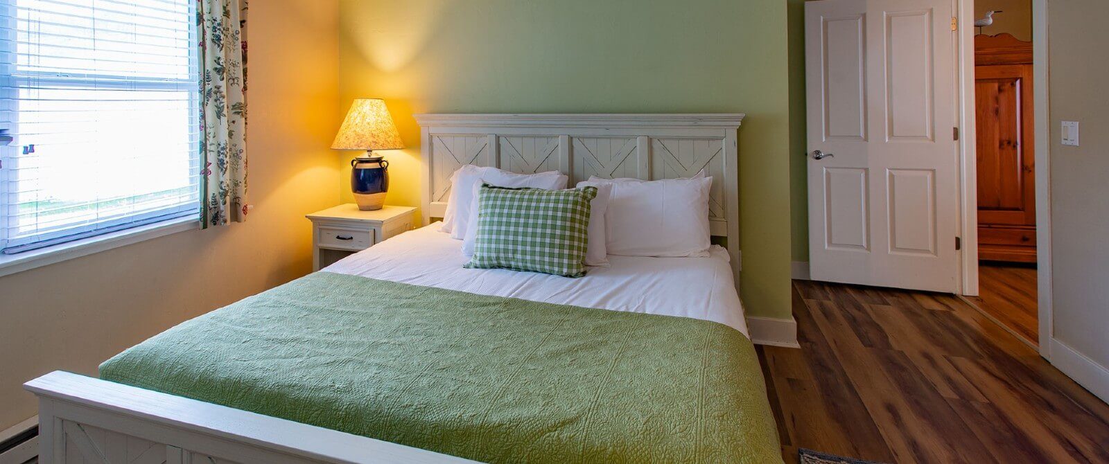Bedroom with queen bed in white and green, table with lamp, a bright window and hardwood floors