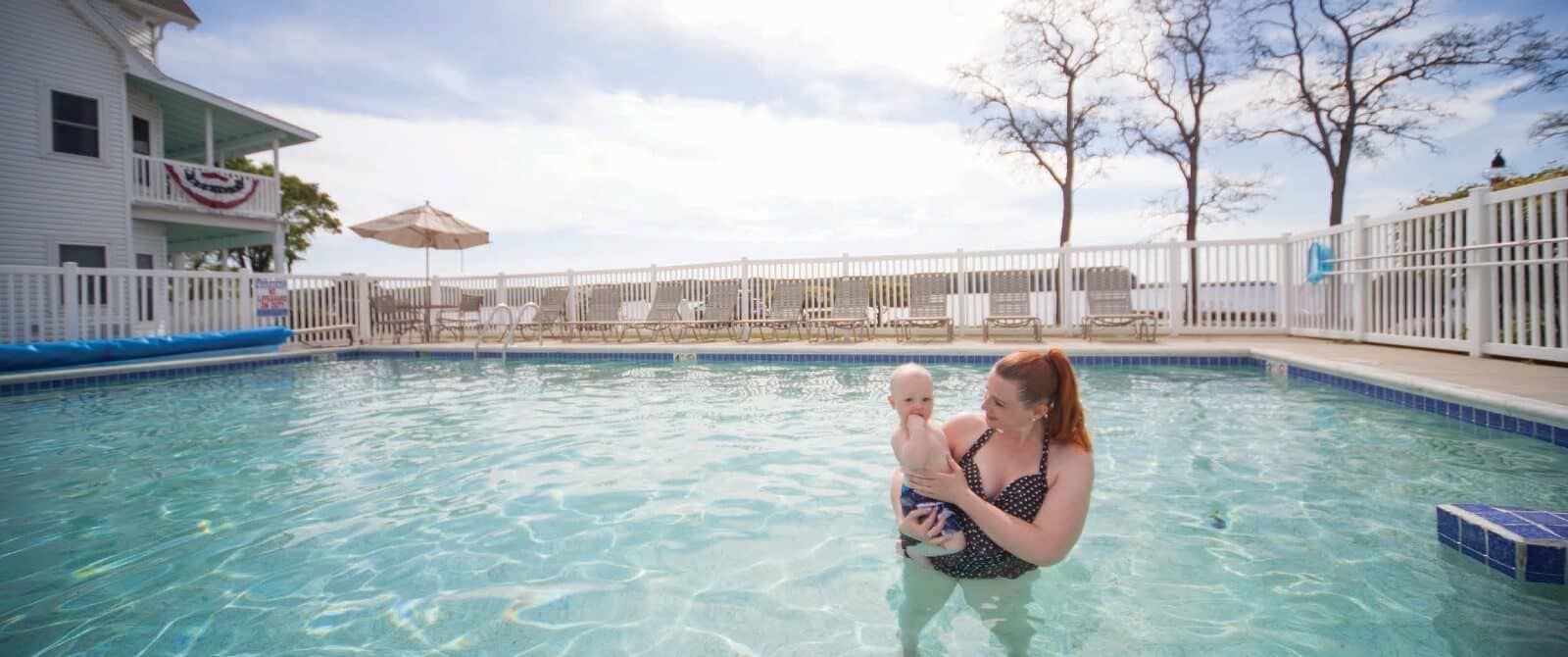 A woman holding a baby in a large outdoor pool next to a hotel building overlooking a lake