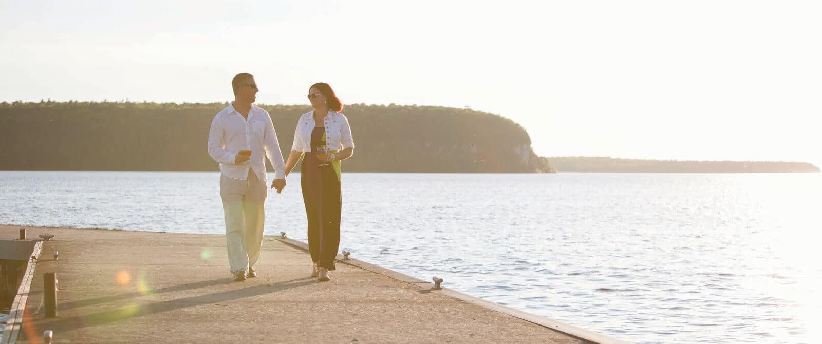 A man and woman holding hands walking on a paved dock near a large lake