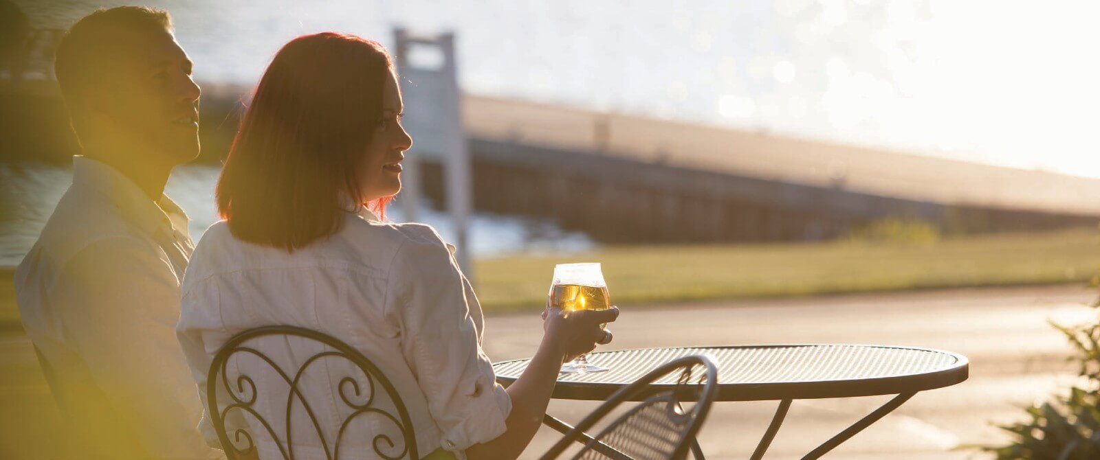 A man and woman, holding a glass of wine, sitting at a wrought iron table outside by a lake