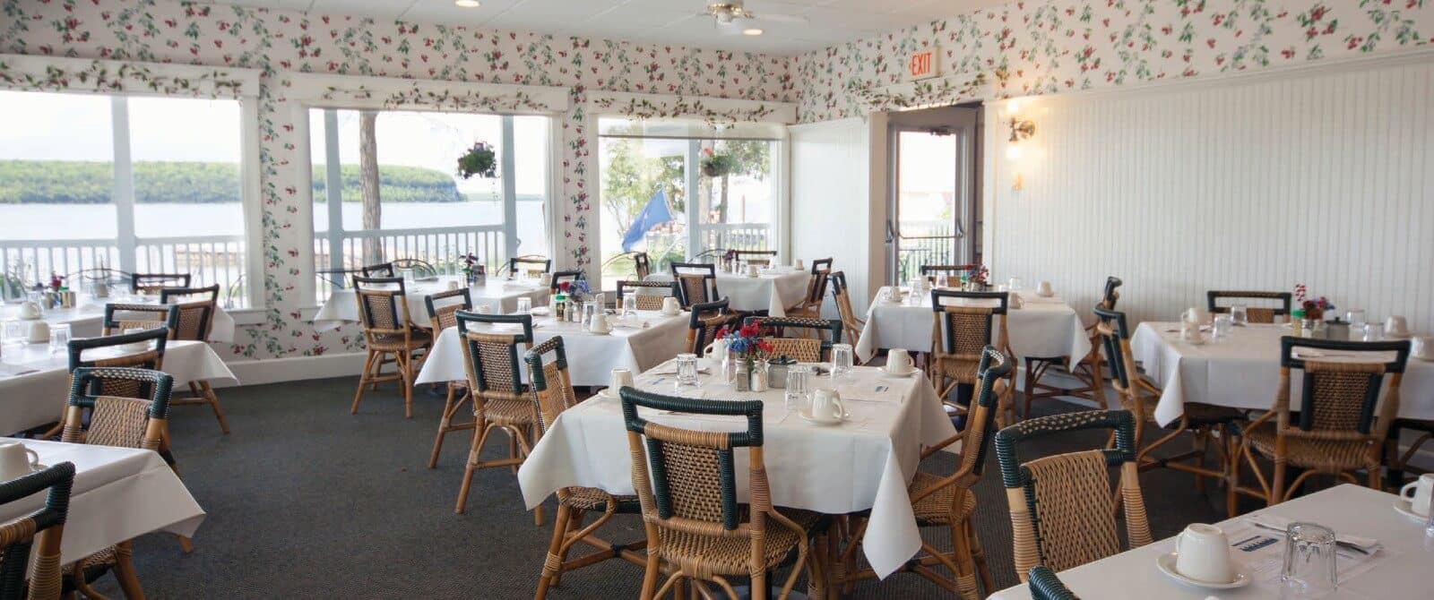 Large dining room with a wall of windows, floral wallpaper and ten tables each with white tablecloths and four chairs