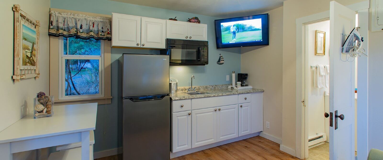 Interior cottage kitchen with white cabinets, fridge, corner mounted TV and doorway into a bathroom