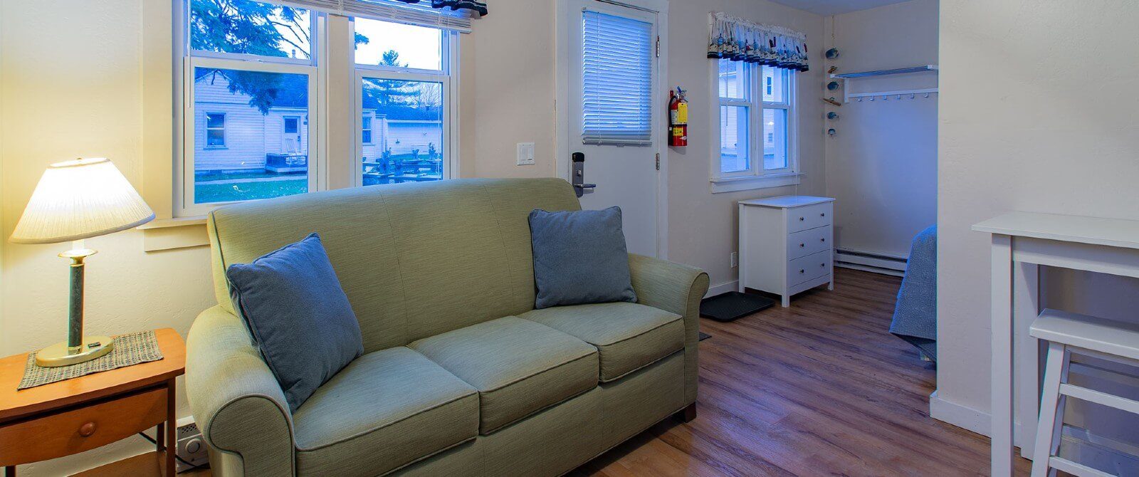 Interior of a small cottage showing couch in living room, side table with lamp and side area with bed and dresser