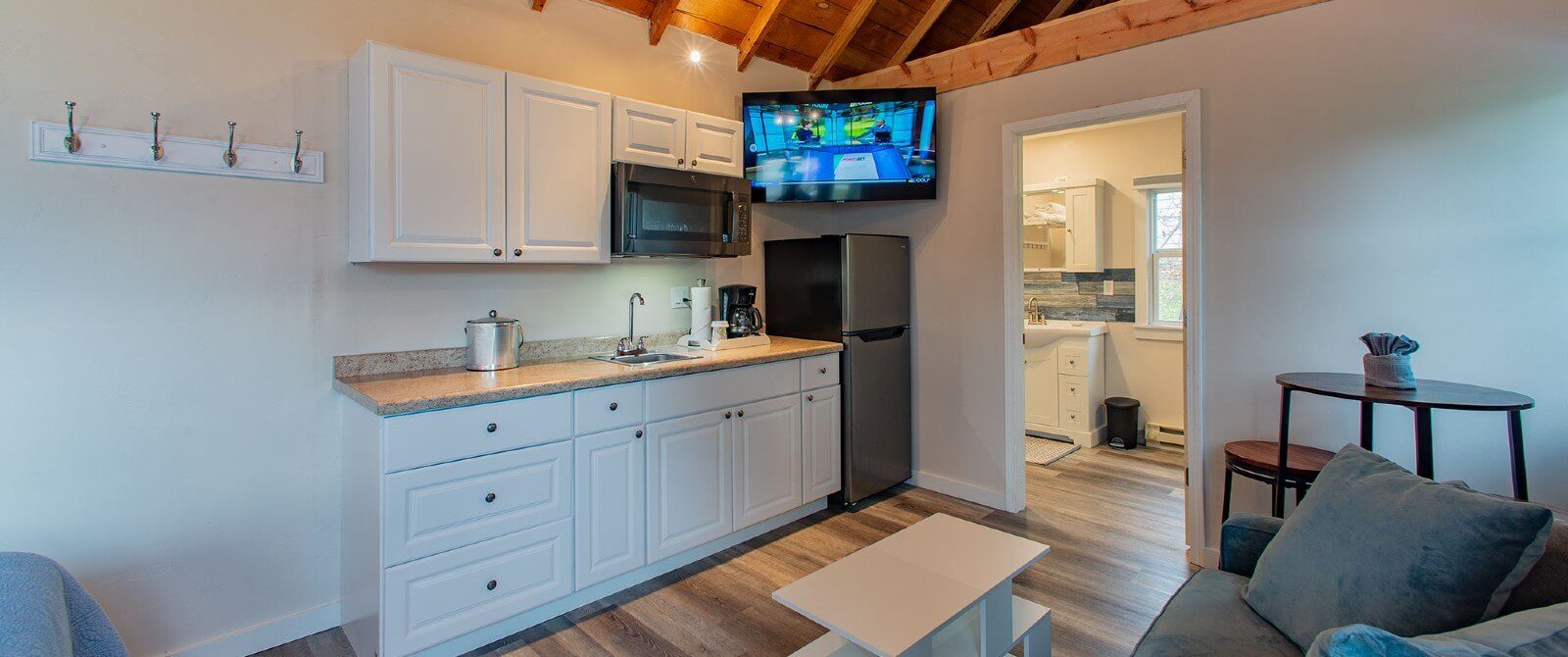 Interior of a cottage with white kitchenette, couch, hardwood floors and doorway into a bathroom