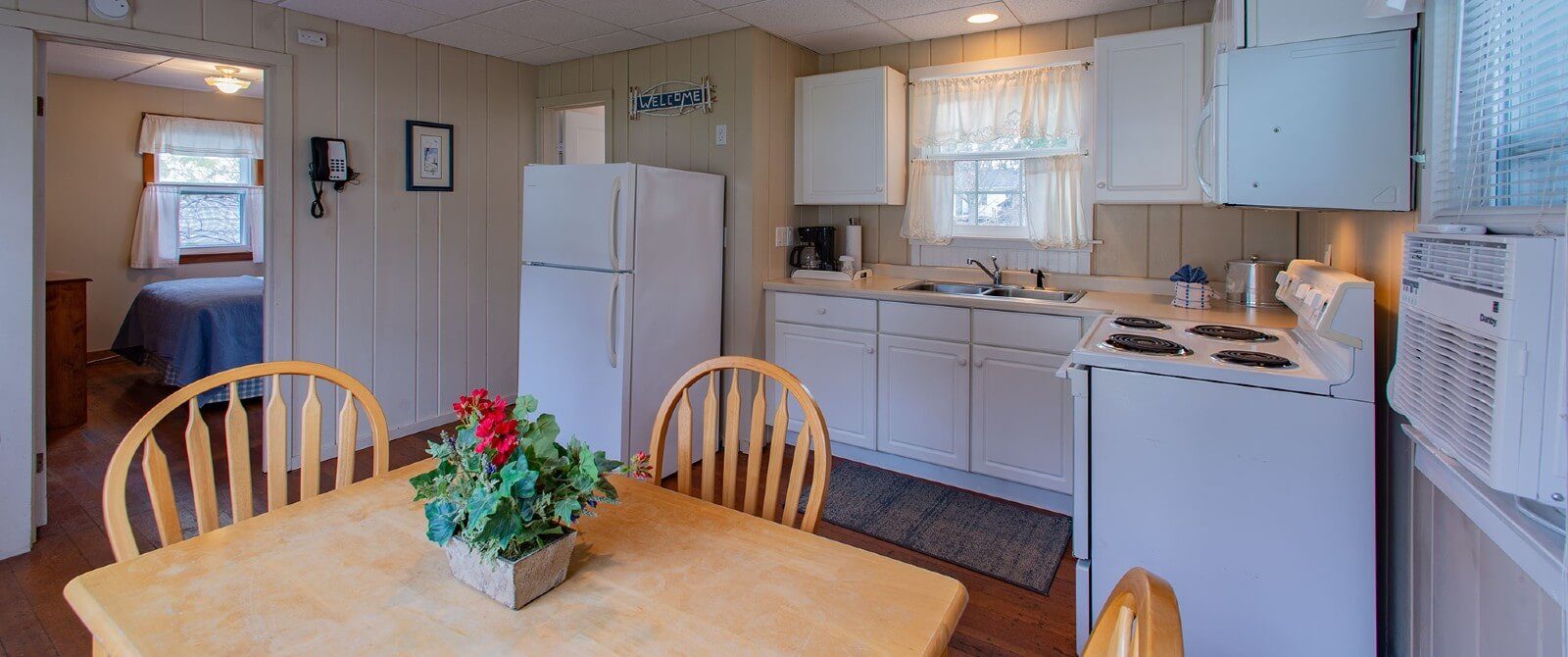 Interior of a cottage with a small white kitchen, wood table with four chairs and doorway into a bedroom