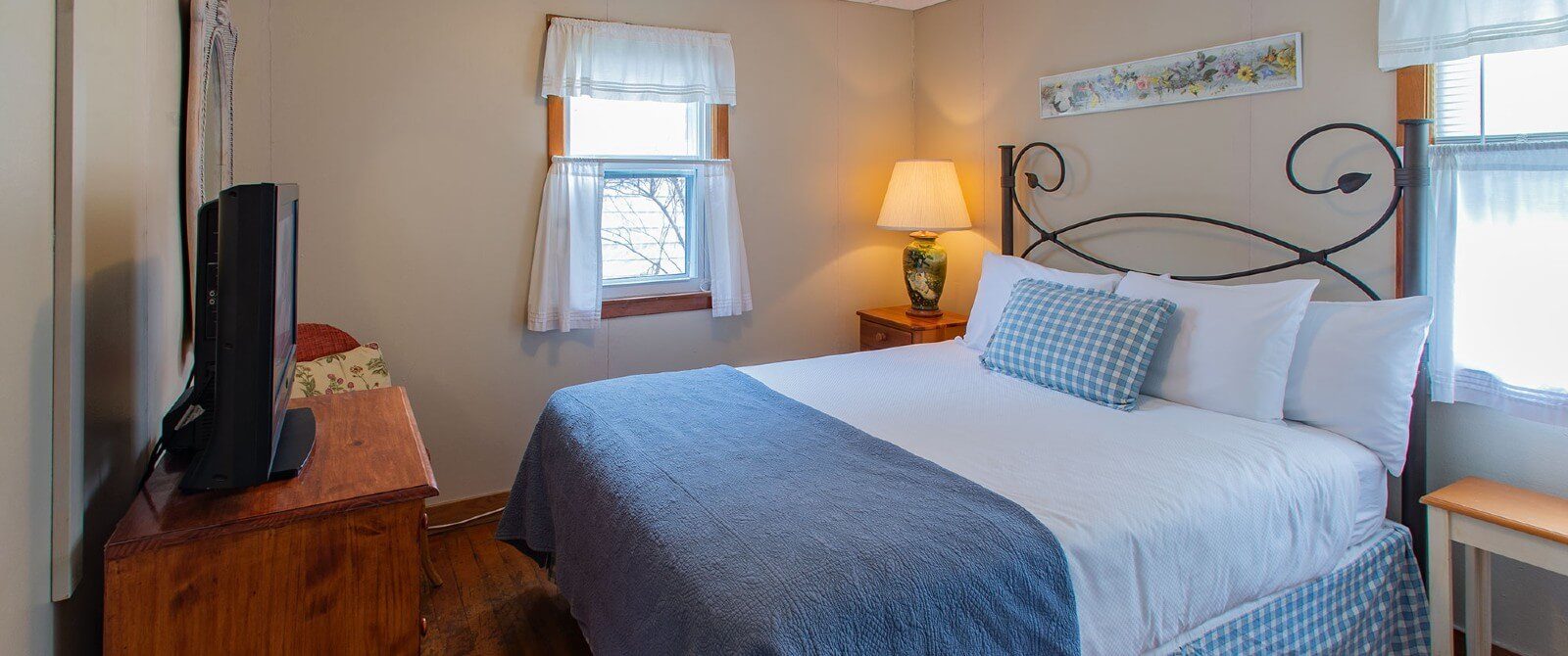 Small bedroom with queen bed in white and blue, dresser with TV and two windows with white curtains