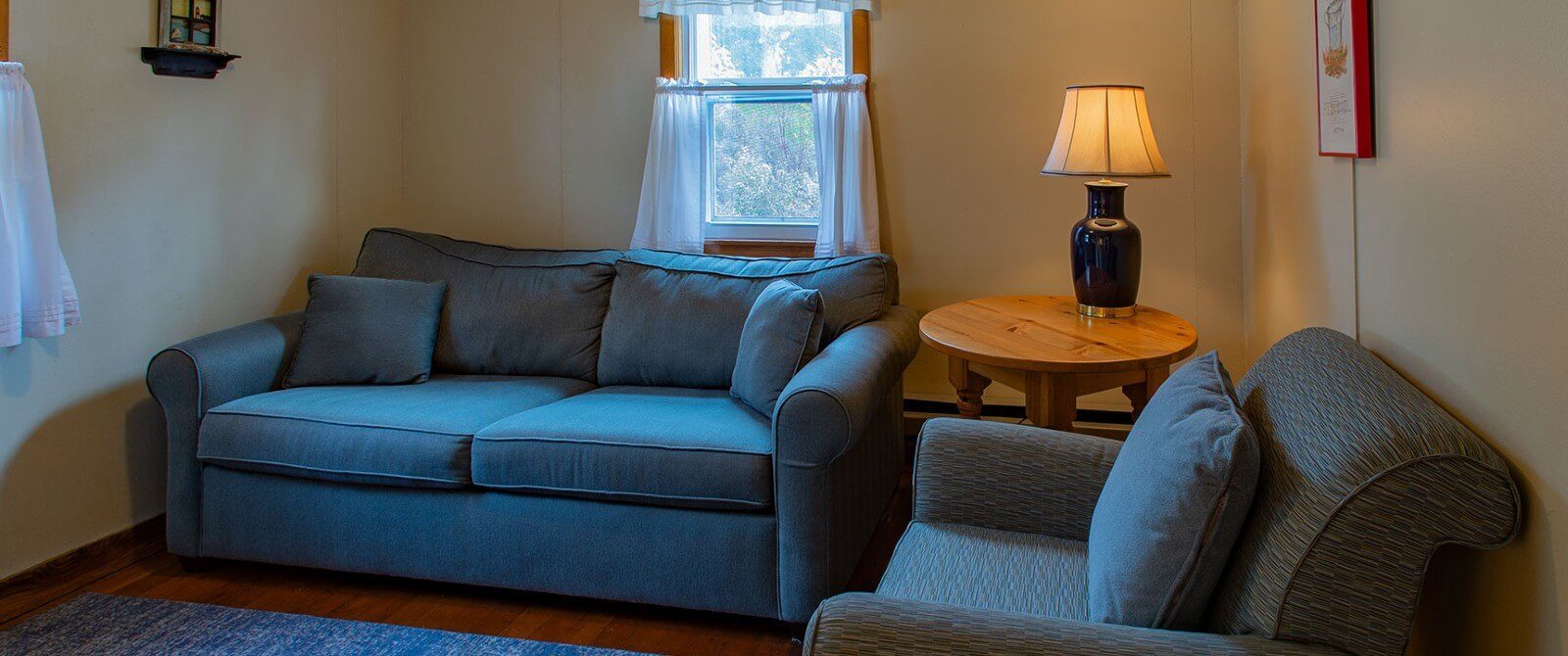 Living room with a couch, chair, round table with lamp, rug and windows with white curtains