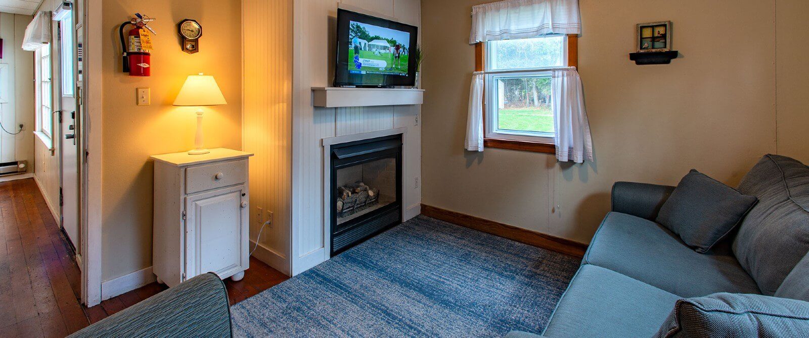 Cottage living room with plush couch, fireplace with TV above, table with lamp and window with white curtains