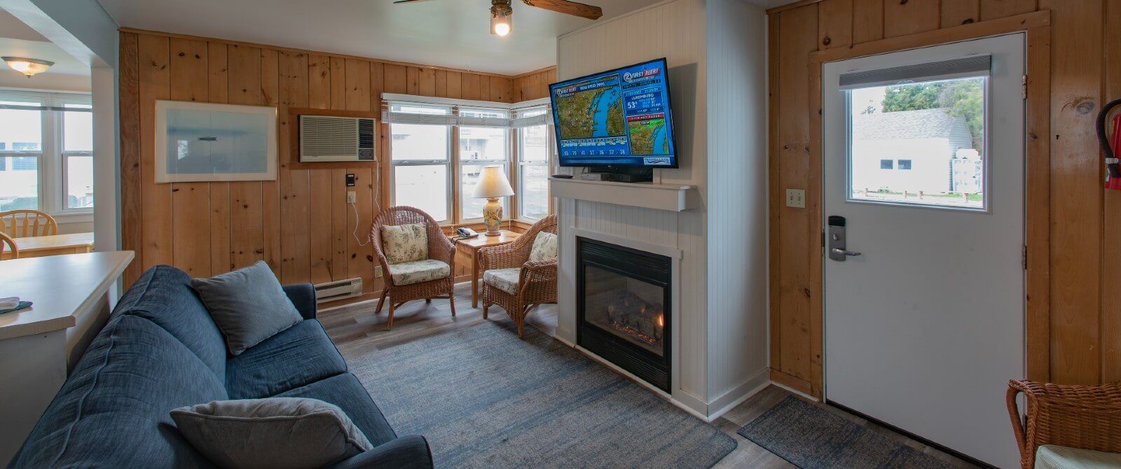 Interior of a cottage with living room, couch, wicker sitting chairs, gas fireplace with flat screen TV and wood paneled walls