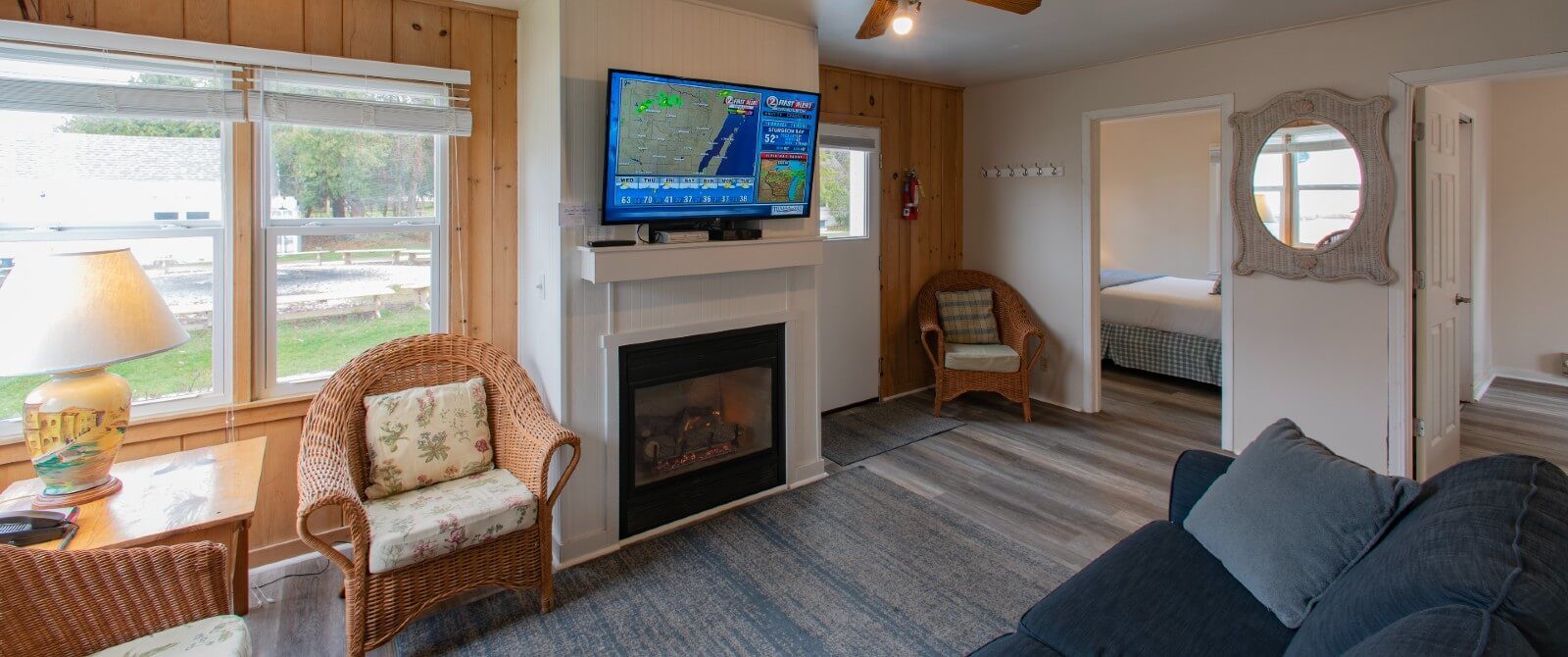 Interior of a cottage with living room, couch, wicker chairs, gas fireplace with TV and doorway into a bedroom