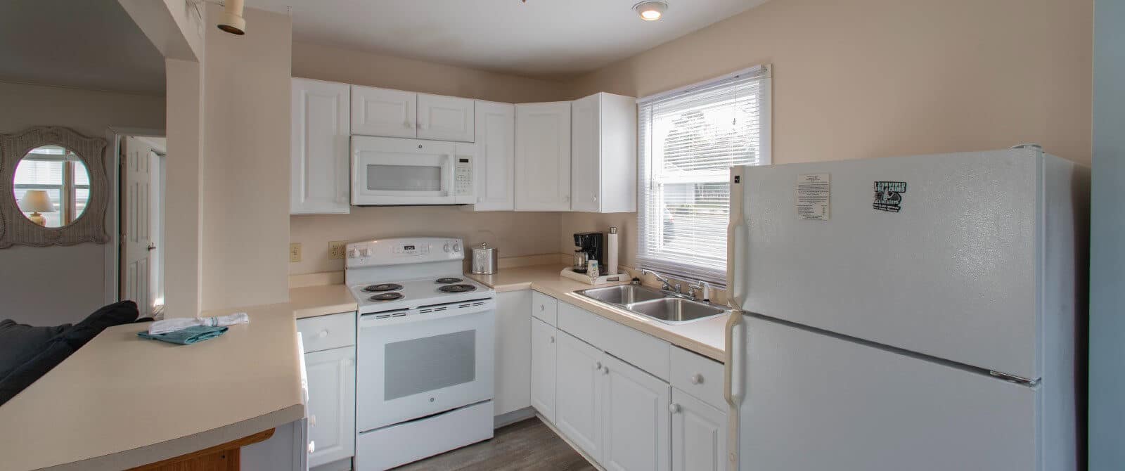 Small white kitchen with cream countertops and large window with blinds