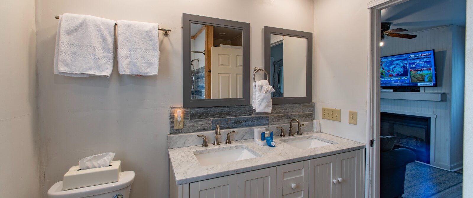 Elegant bathroom with double sink vanity, two framed mirrors, white hanging towels and doorway into a living room