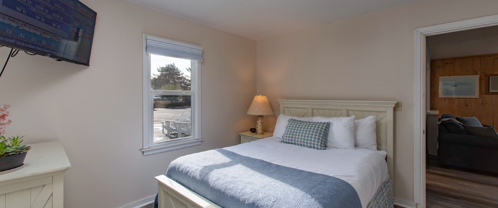 Small bedroom with queen bed, single window, corner hung TV and doorway into a living room