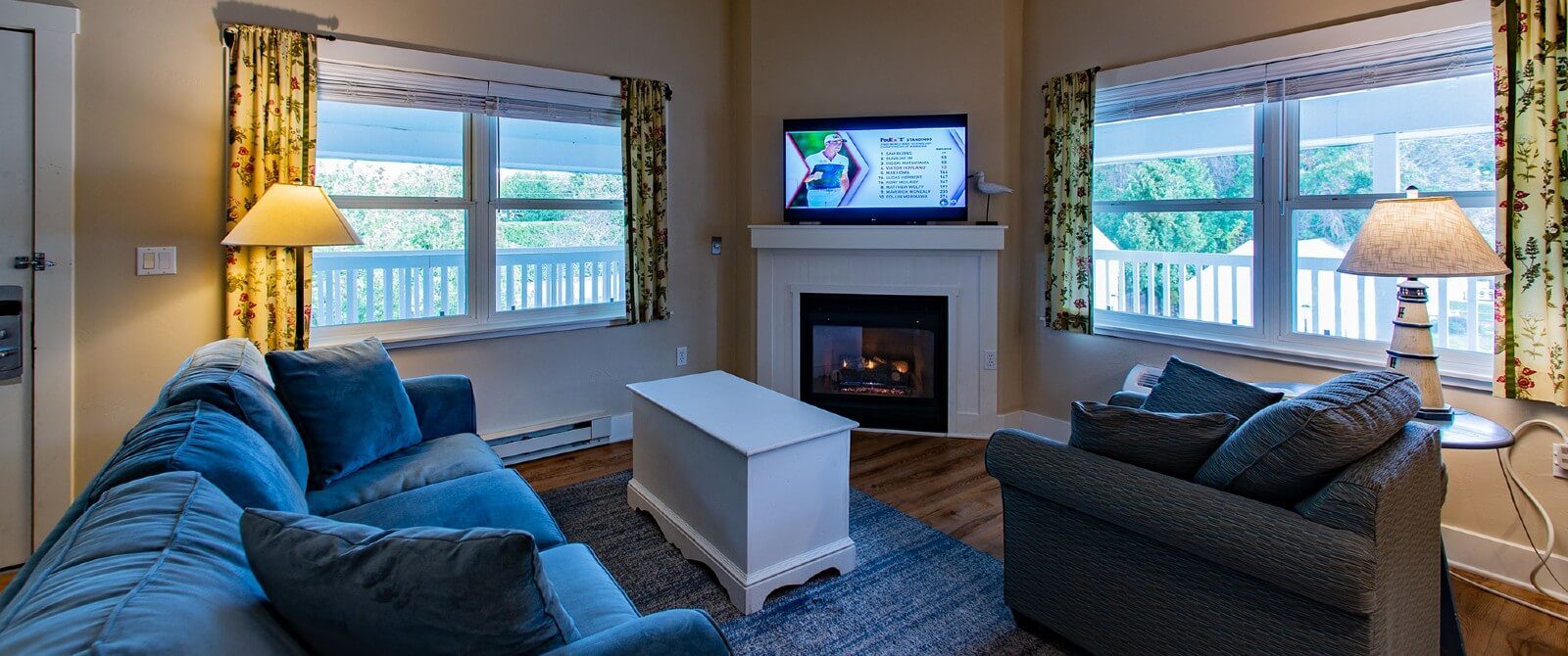 Living room with couch, chair, coffee table in front of gas fireplace with large windows with floral curtains