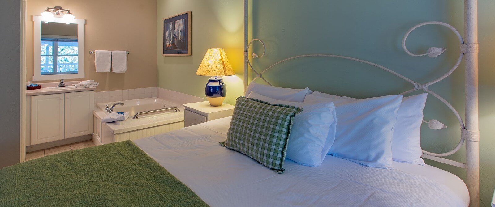 Bedroom with queen bed in green and white linens, vanity with large mirror and corner jacuzzi tub