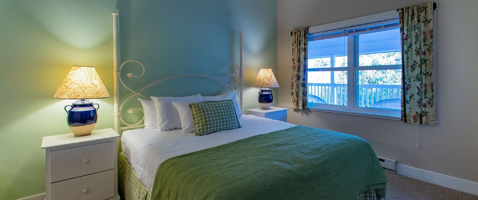 Small bedroom with queen bed in green and white blankets, two side tables with lamps and large window