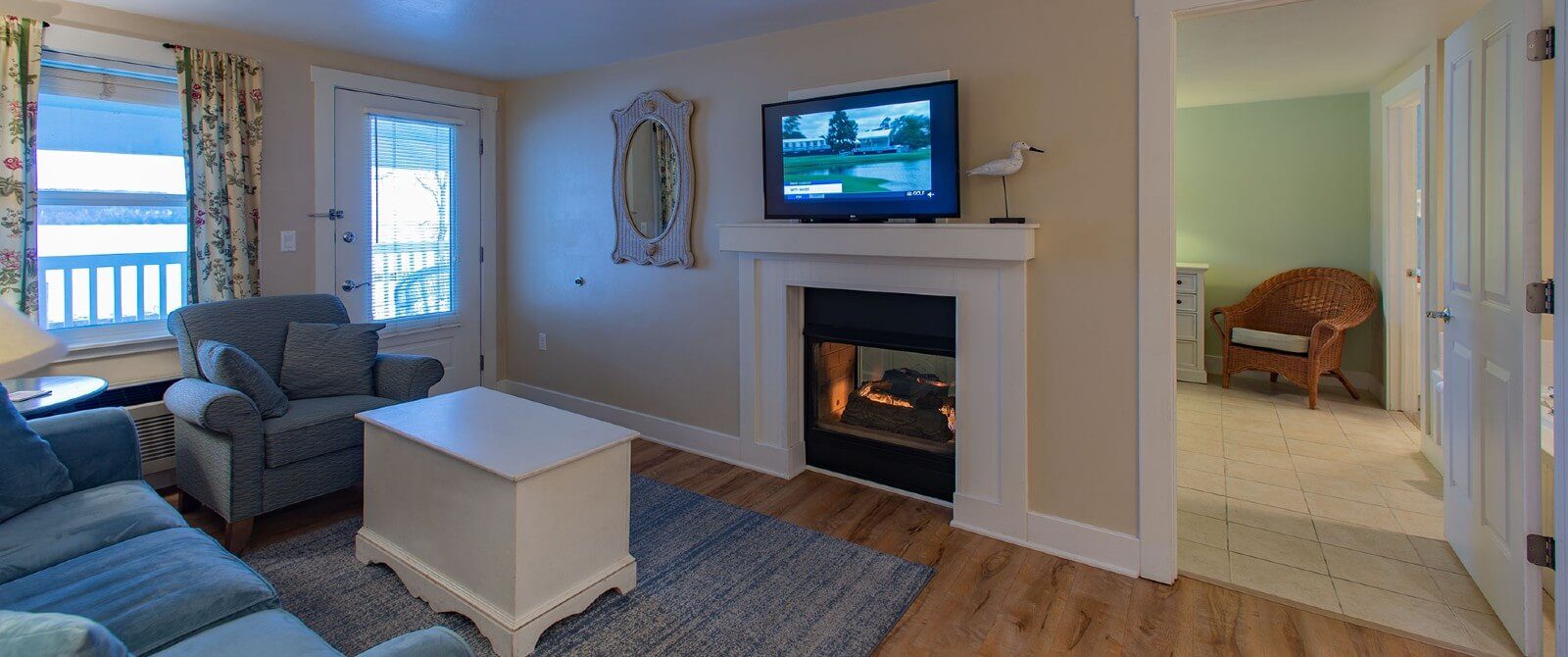 Living room with couch, chair and white coffee table in front of a gas fireplace with doorway into a bedroom