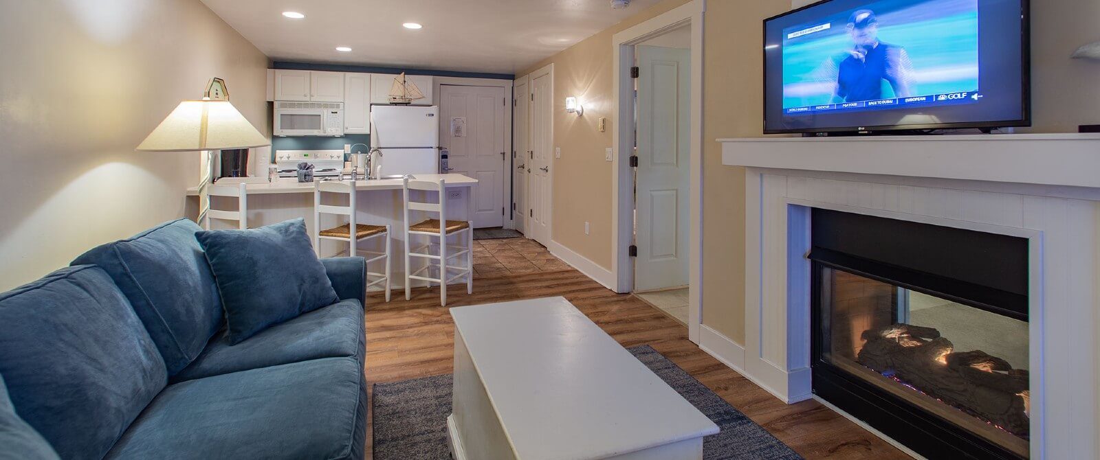 Cozy living space with kitchenette, plush blue couch and coffee table in front of gas fireplace with TV above