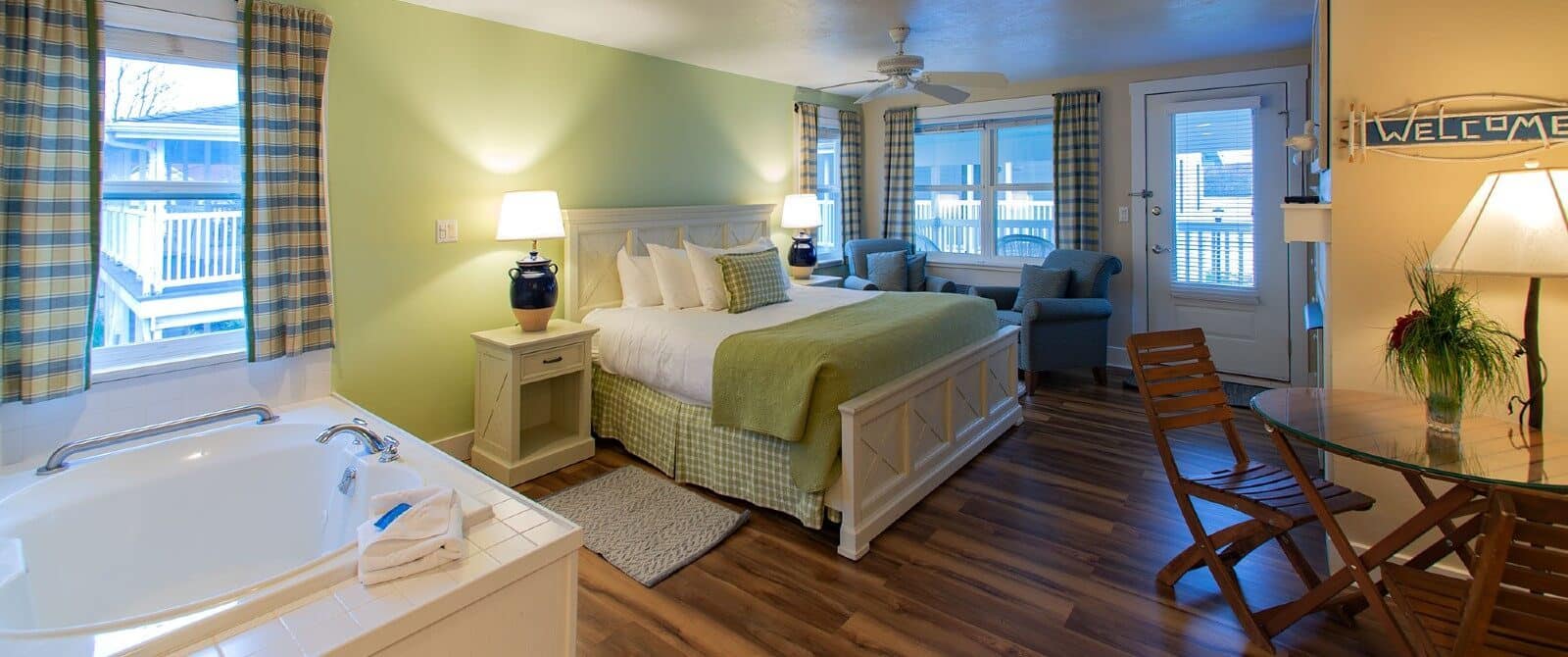Bedroom with hardwood floors. queen bed, jacuzzi tub, sitting chairs, large windows with plaid curtains