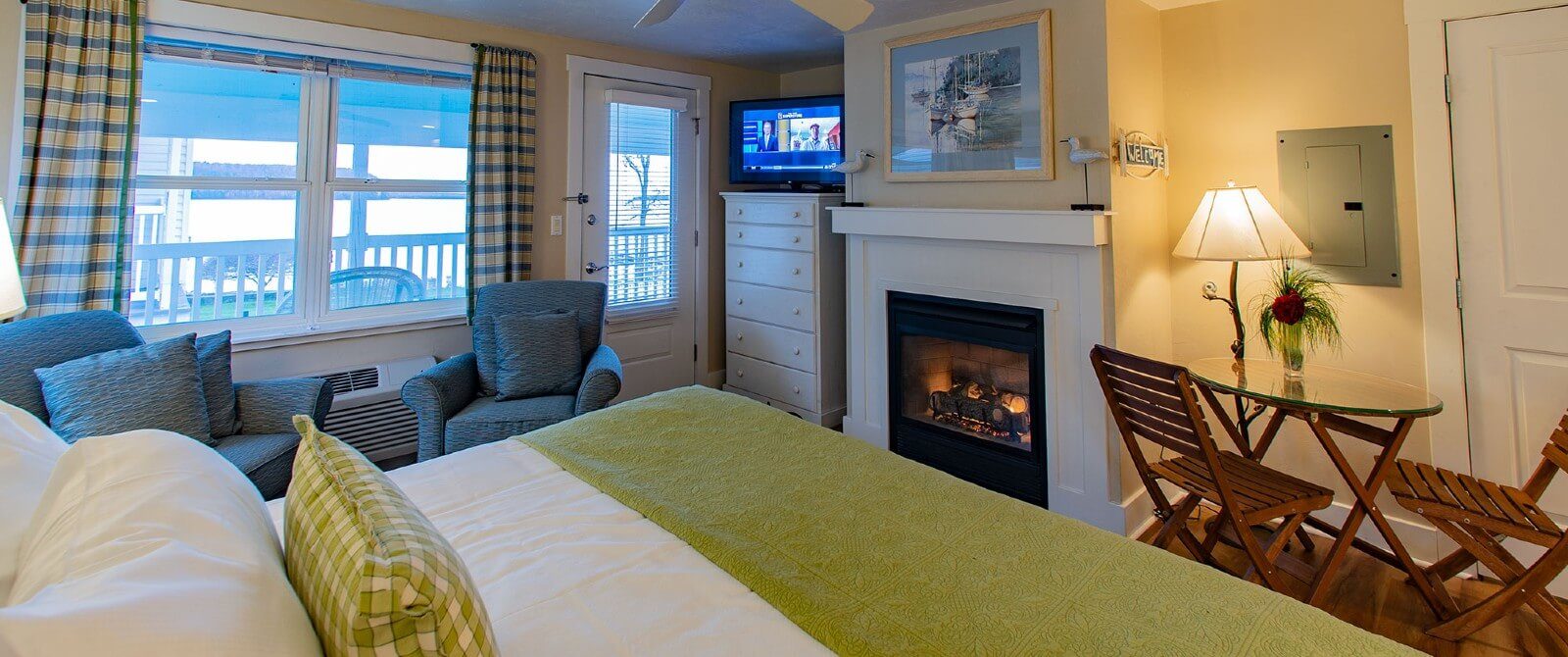 Bedroom with queen bed, sitting chairs, large windows with plaid curtains, gas fireplace and dresser with TV