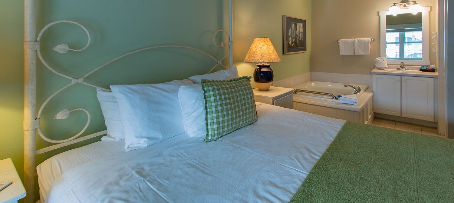 Bedroom with queen bed in green and white linens, corner jacuzzi tub and vanity with white framed mirror