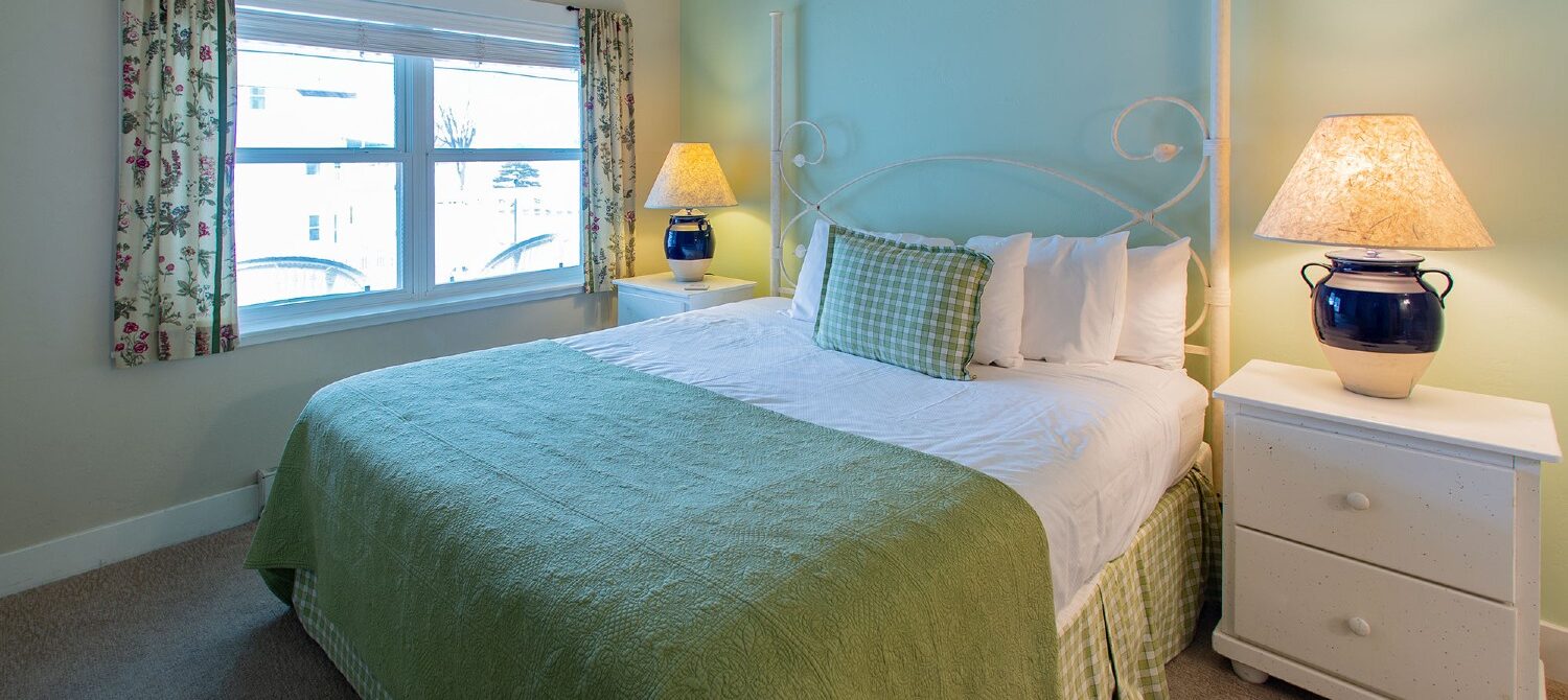 Bedroom with queen bed, white side tables with blue lamps and large picture window overlooking the water