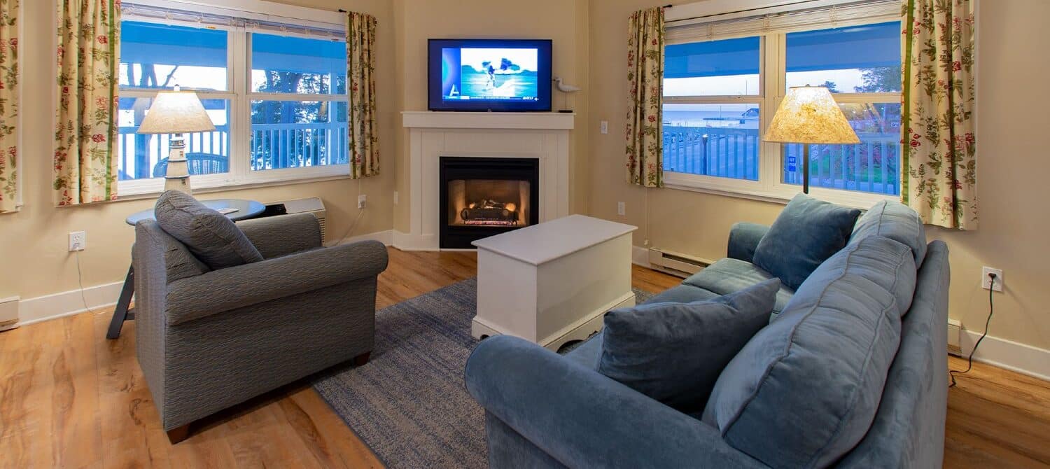 Living room with plush couch and chair, table, gas fireplace with TV and two large windows overlooking the water
