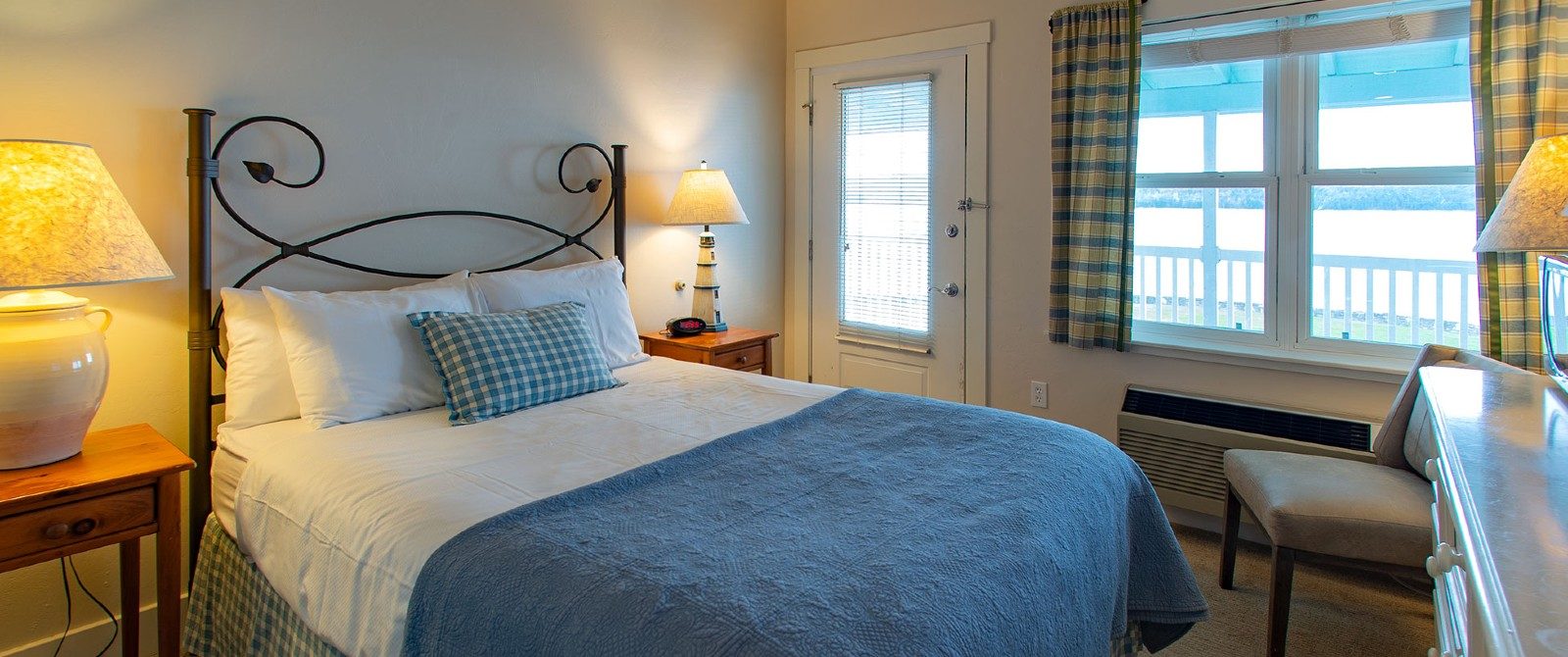 Cozy bedroom with queen bed, side tables with lamps and large window with plaid curtains overlooking the water