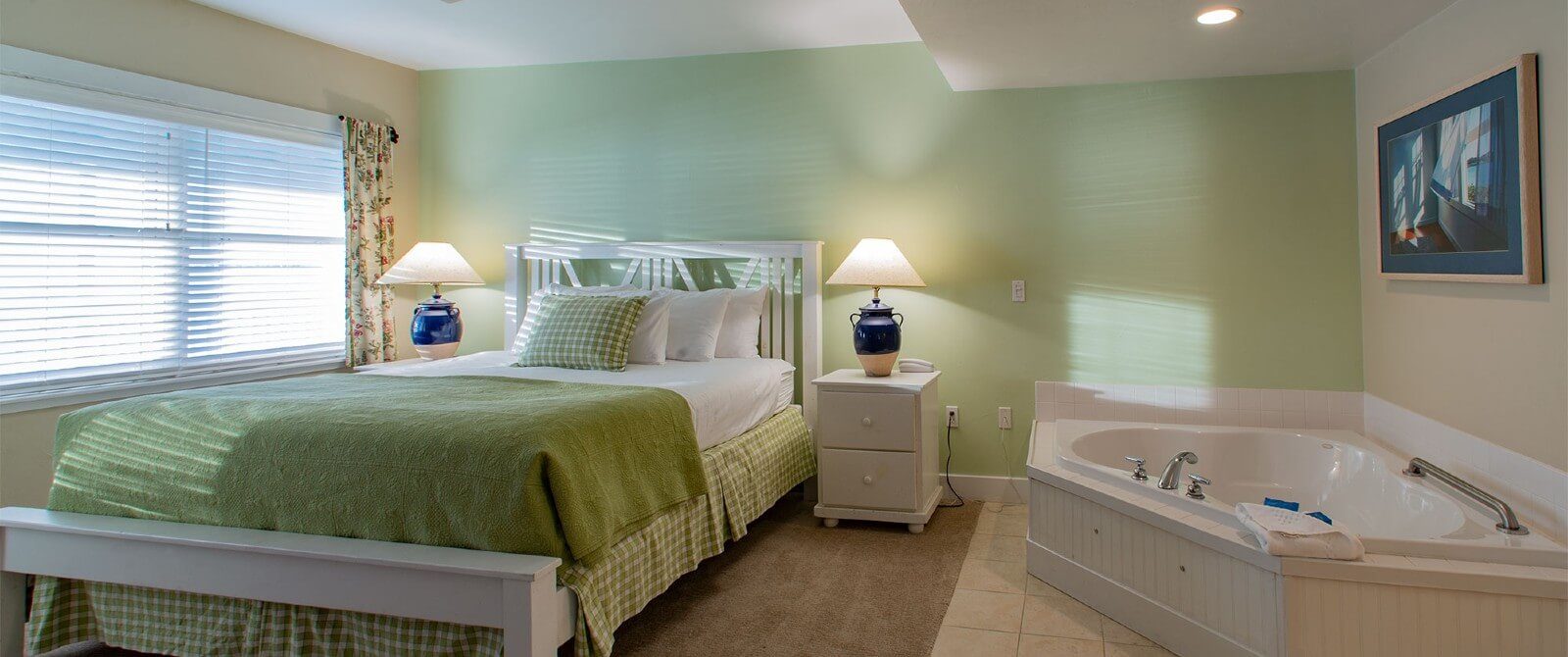 Elegant bedroom in hues of green and white with bed near large window with blinds and corner jacuzzi tub