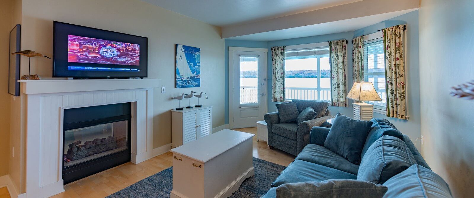 Hotel suite living room with plush blue couch and chair, coffee table in front of a gas fireplace, TV and large bay window overlooking the water