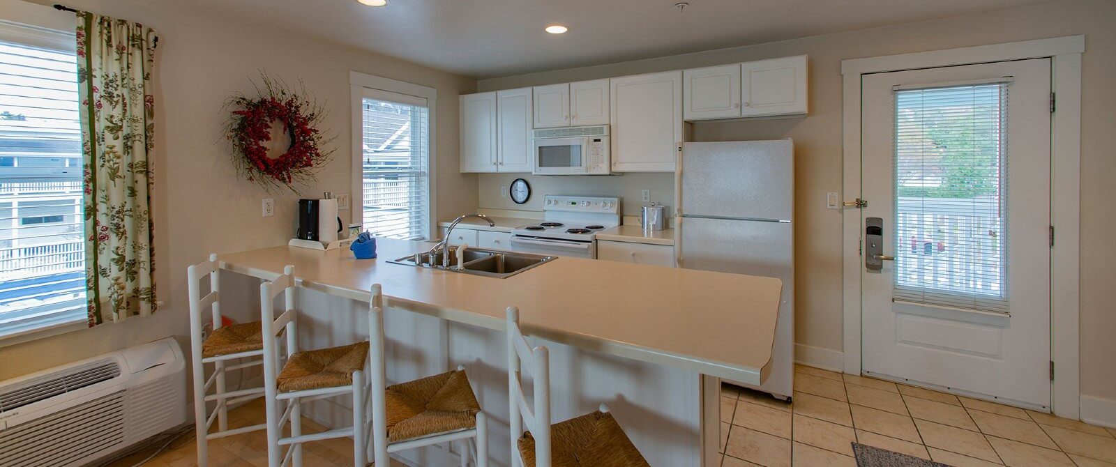 Small kitchen with tiled floor, white cabinets, four stools by the island and windows with blinds and floral curtains