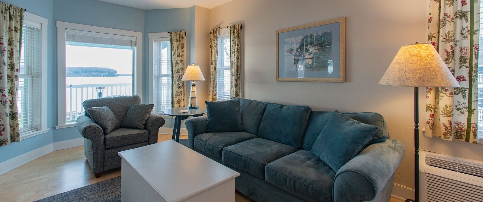 Living room with plush couch and chair, white coffee table and two lamps near bay window overlooking water