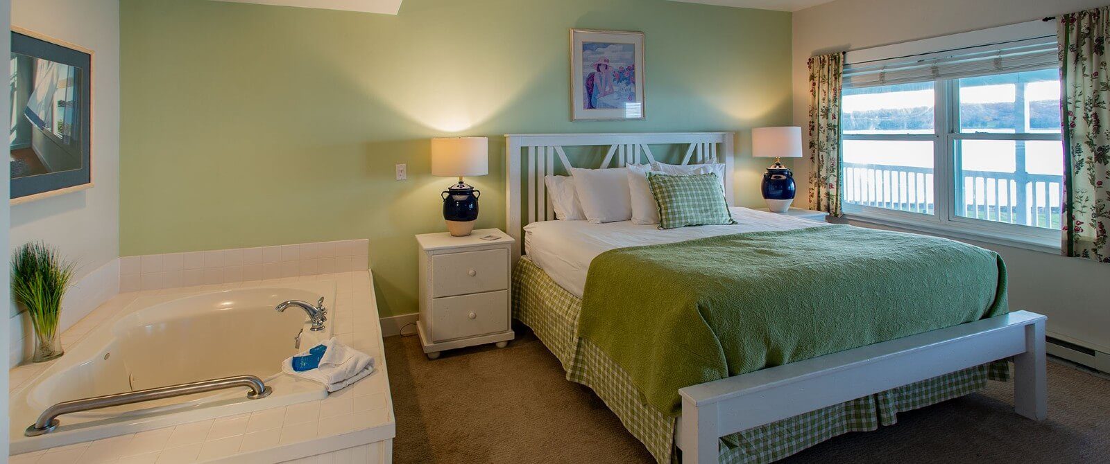 Cozy bedroom with bed in green and white linens near window overlooking the water and a large jacuzzi tub