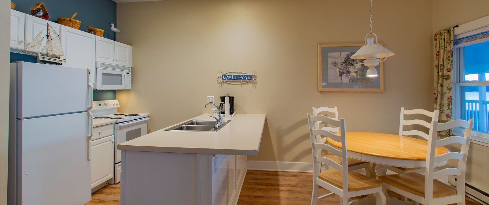 Small white kitchen with sink island and round table with four chairs by a large window with floral curtains