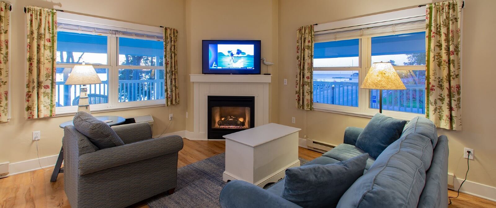 Living room with plush couch and chair by a corner gas fierplace flanked by large windows with floral curtains