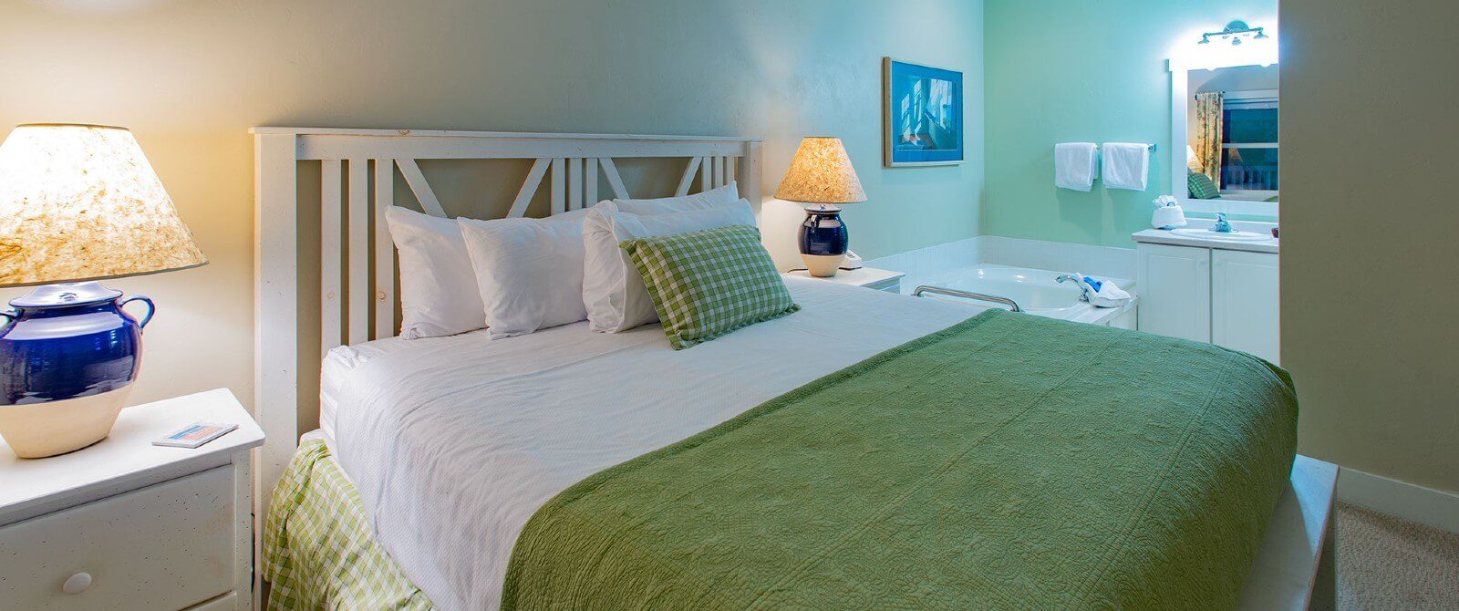 Bedroom with bed in green and white linens, side tables with lamps and corner jacuzzi tub near vanity with mirror