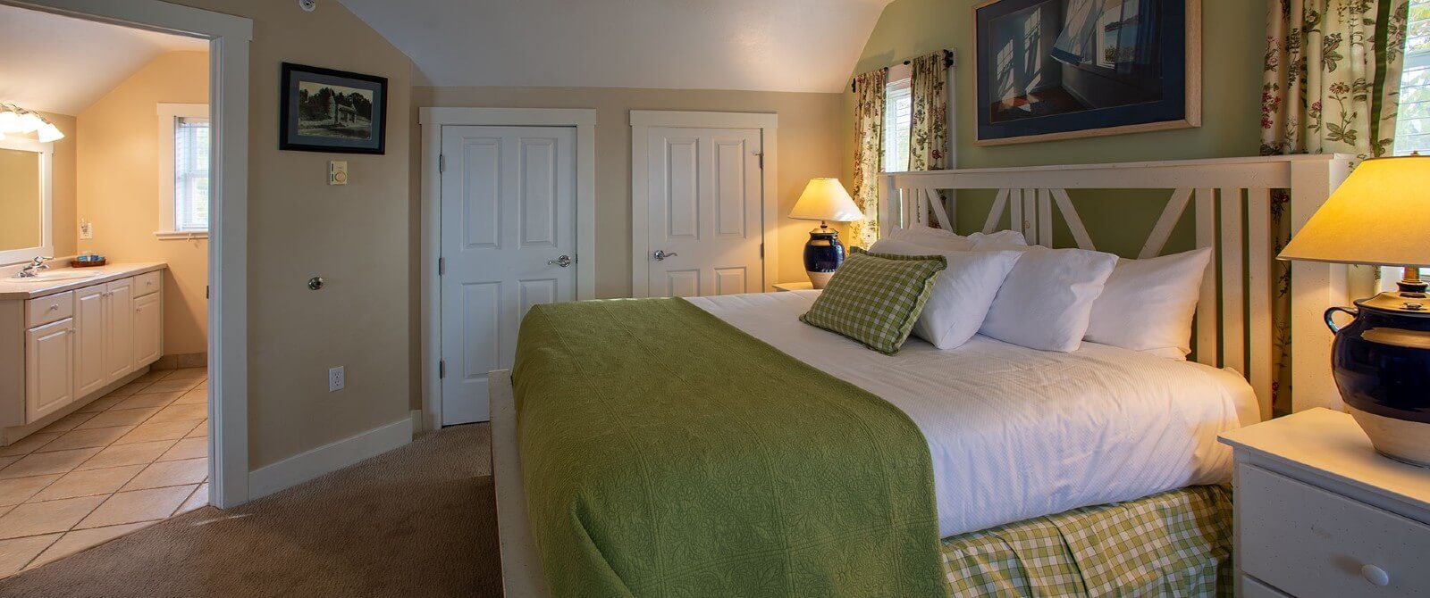 Bedroom with king bed in white and green linens, side tables with lamps and doorway into a large bathroom